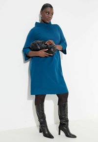 Teal ylisuurta mekkoa, jossa on suppilokaulus ja kolmannen neljänneksen hihat; paritettuna mustan clutch-laukun ja polveen asti ulottuvien mustien korkokenkien kanssa.