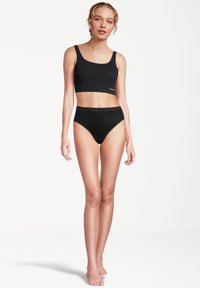 Junge Frau, die barfuß steht und ein schwarzes Crop-Top und schwarze High-Waist-Unterhosen trägt, vor einem schlichten weißen Hintergrund.