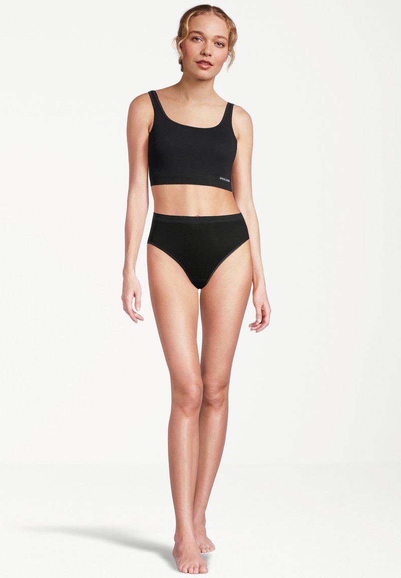 Junge Frau, die barfuß steht und ein schwarzes Crop-Top und schwarze High-Waist-Unterhosen trägt, vor einem schlichten weißen Hintergrund.