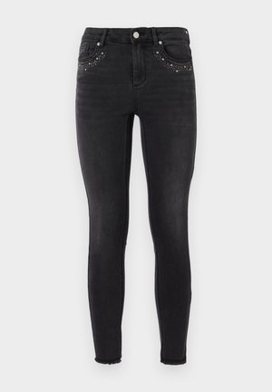Black skinny jeans made of denim with a slight sheen. Features studded embellishments on front pockets and a frayed hem at the ankles.