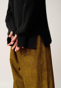 Czarny sweter dziergany z prążkowaną teksturą, zestawiony z szerokimi spodniami w poziome paski w musztardowym kolorze. Sweter ma boczne rozcięcia; ręce złożone razem.