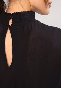 Schwarze Bluse mit hohem, gerafftem Kragen, Schlüsselöffnungsdetail auf der Rückseite und glatter Stoffoberfläche mit subtiler Schimmerwirkung. Zwei Knöpfe am Halsabschluss.