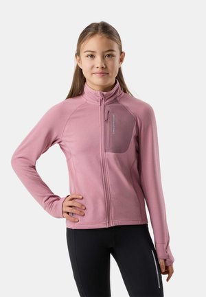 Ung person iført en tætsiddende lyserød lynlåstrøje med brystlomme og sorte leggings, stående med den ene hånd på hoften mod en ensfarvet baggrund.