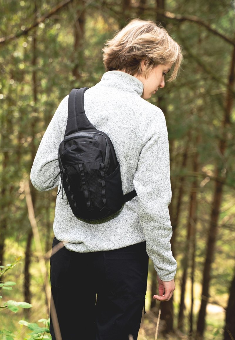 Zaino a sacca nero con superficie strutturata, tracolla regolabile e accenti elastici. Maglione grigio in pile e pantaloni scuri in un ambiente boschivo.