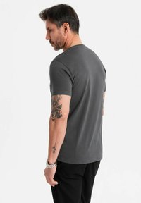 Šedé bavlněné tričko s krátkým rukávem a kulatým výstřihem, prezentované na mužském modelu. Výrazný tetování na levém rameni.