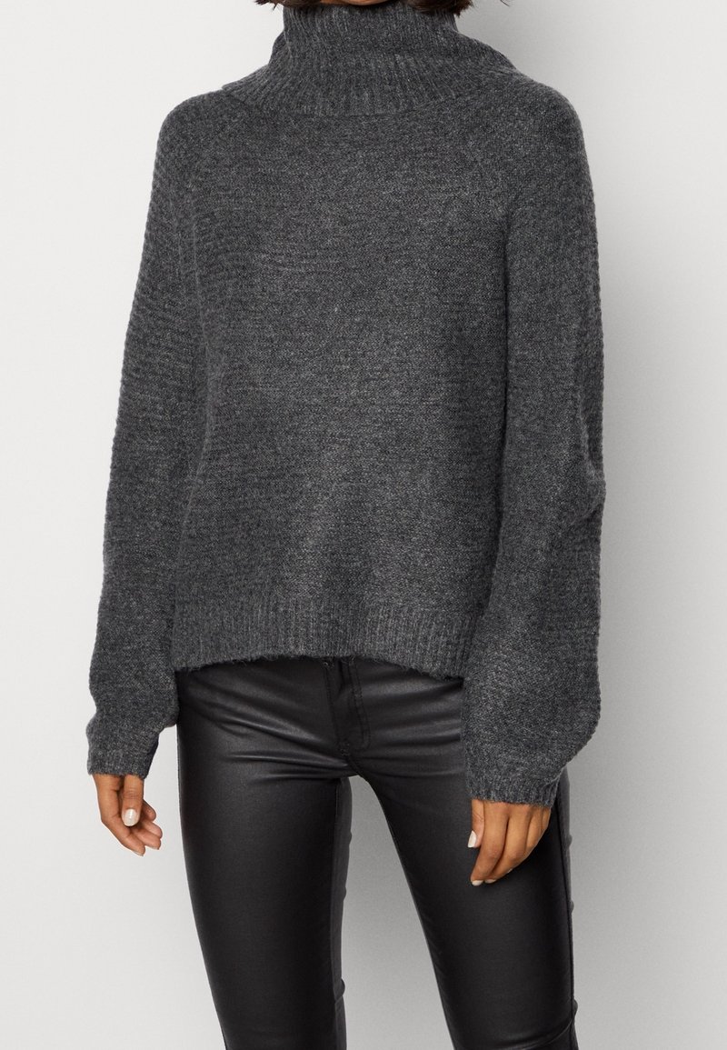 Femme portant un pull à col roulé en grosse maille gris foncé et un pantalon en similicuir noir, debout devant un fond blanc uni.