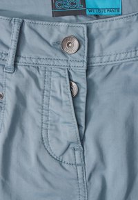 Helle blaue Baumwollhose mit Reißverschluss und Metallknopfverschluss, ausgestattet mit Taschen und sichtbaren Steppnähten. Markenlabel innen im Bund eingestickt.