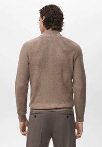 Uomo con capelli ricci che indossa un maglione marrone a coste e pantaloni marroni, in piedi di spalle contro uno sfondo bianco uniforme.