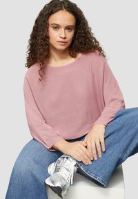 Rosa strukturierte Langarmbluse mit weitem Ausschnitt, kombiniert mit blauen weiten Jeans und silbernen Sneakers, sitzt auf einem weißen Würfel.