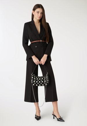 Abito strutturato nero con pantaloni a gamba larga, cintura in pelle marrone e borsa nera decorata con borchie argento. Décolleté nere con punta affilata.