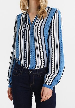 Frau trägt eine langärmlige Bluse mit vertikalen blauen, schwarzen und weißen Streifen und Mustern, kombiniert mit dunkelblauen Jeans.