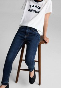 Camiseta blanca de manga corta con "PARIS AMOUR" en letras negras, combinada con jeans de mezclilla azul oscuro y zapatos planos negros sobre un taburete de madera.