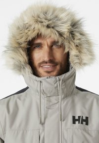 Grijze buitenjas met een faux fur-gevoerde capuchon, zwarte accenten en een logo. Heeft een hoge kraag en ritszakken aan de voorkant.