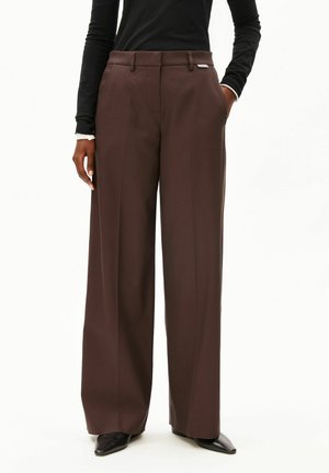 Person trägt hoch taillierte, weit geschnittene braune Hose mit der Hand in der Tasche, kombiniert mit schwarzen spitzen Schuhen und einem schwarzen Langarmoberteil.