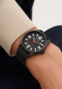 Black analog watch with a textured dial, large white numbers, and a red accent. Features a rubber strap with a distinctive pattern.