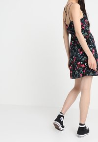 Czarna sukienka w kwiaty z cienkimi ramiączkami, zdobiona różowymi i zielonymi tropikalnymi wzorami. Połączona z czarnymi niskimi trampkami.