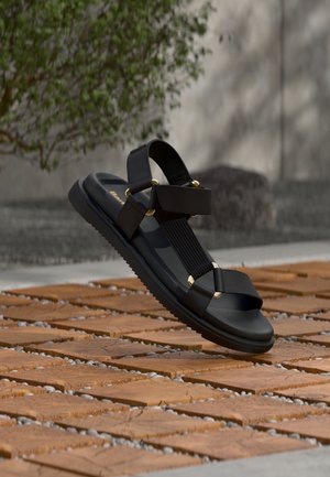 Sandală neagră cu accente de inele aurii plutește deasupra unei suprafețe exterioare din plăci de lemn și pietricele, cu un fundal de copac și piatră neclar.