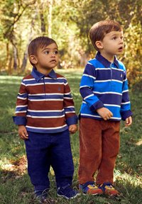 Dos niños llevan camisetas polo de manga larga: una marrón con rayas y la otra azul. Ambos tienen pantalones de pana, uno en azul marino y el otro en óxido.