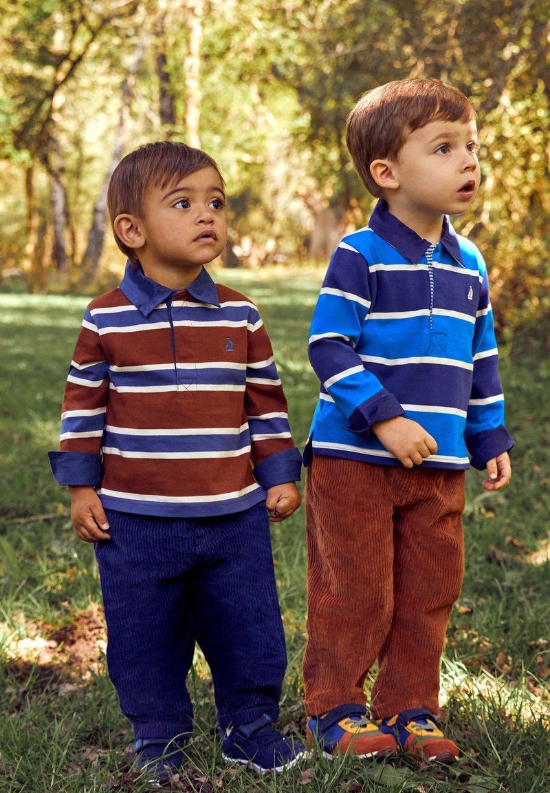 Dos niños llevan camisetas polo de manga larga: una marrón con rayas y la otra azul. Ambos tienen pantalones de pana, uno en azul marino y el otro en óxido.