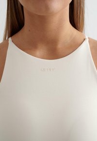 Top sportivo bianco con un collo alto e spalline sottili. Presenta un logo goffrato discreto sul davanti. Texture liscia.