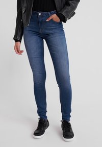 Jeans ajustados en denim azul medio, con un diseño clásico de cinco bolsillos, cintura alta y una textura suave, combinados con zapatillas negras.