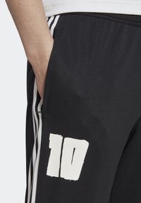 Person som bär svarta träningsbyxor med vita ränder och ett stort vitt nummer "10" på låret, med handen i fickan.