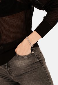 Zilveren kettingarmband met onderling verbonden schakels, gedragen aan een hand die in de zak van zwart denim jeans rust, met een gebreid mouwtrui.