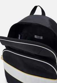 Czarny plecak z główną przegrodą na zamek błyskawiczny, z białymi i żółtymi paskami oraz tkana metka z logo wewnątrz.