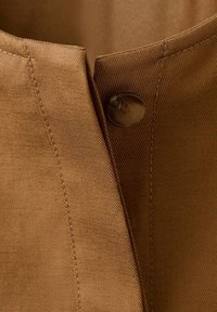 Gros plan sur un tissu marron avec un unique bouton marron foncé cousu près d'une couture verticale et d'un bord plié.