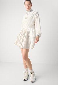 Vit plisserad klänning med hög krage och elastisk midja. Långa ärmar, mjukt material, kombinerad med sportiga mångfärgade sneakers.