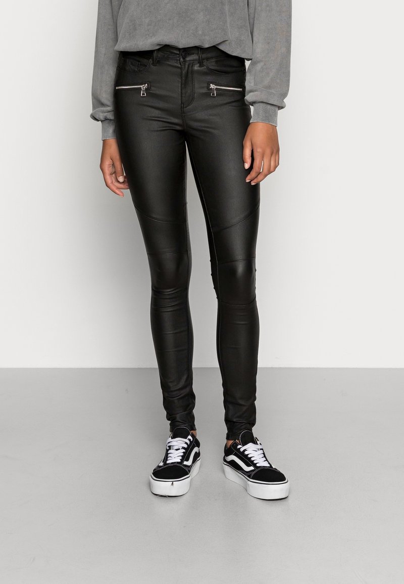 Personne portant un pantalon en cuir noir avec des poches zippées, un haut gris à manches longues et des baskets noires et blanches à lacets, debout sur un sol gris.