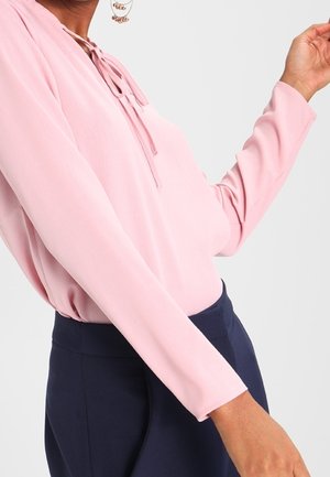 Top a maniche lunghe in tessuto rosa chiaro con scollo regolabile. Abbinato a pantaloni blu navy, che mettono in risalto una texture liscia.