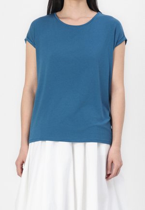 Vrouw draagt een blauw T-shirt met korte mouwen en een witte rok, staand tegen een effen lichte achtergrond.