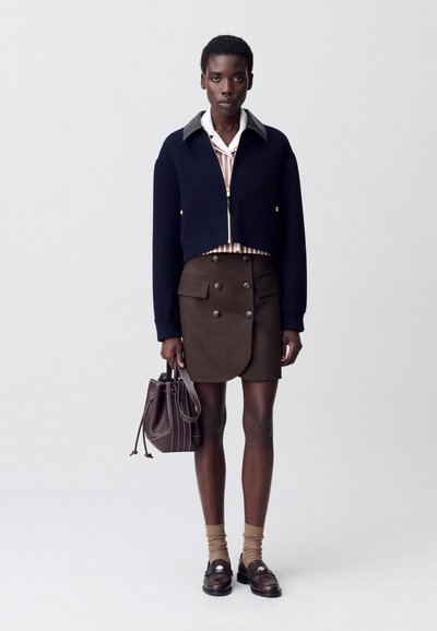 Persona che indossa una giacca blu navy corta, una camicia a righe, una gonna marrone con bottoni, calze beige, mocassini scuri e tiene una borsa in pelle marrone.