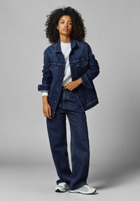 Giacca di jeans e pantaloni a gamba larga in blu scuro con cuciture chiare. Strato sopra una t-shirt bianca. Calzature comprendono sneakers grigie.
