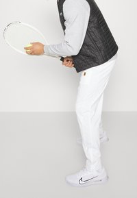 Vita tennisbyxor, grå sweatshirt och svart vadderad väst. Håller en tennisracket med en gul tennisboll. Vita skor med en svart logotyp.