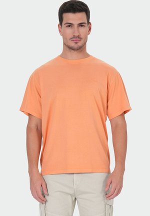 Man wearing plain orange short-sleeve t-shirt and light beige cargo pants standing against a plain background.