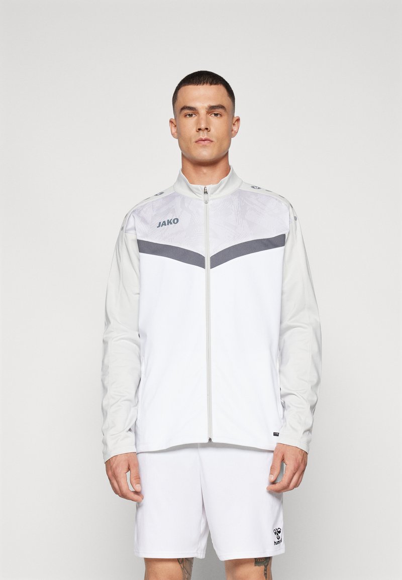 Jachetă sportivă albă cu panouri gri, cu fermoar și logo-ul "JAKO" pe piept. Însoțită de pantaloni scurți albi. Textură fină.