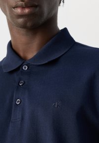 Artimas asmens kadras, dėvinčio tamsiai mėlyną Calvin Klein polo marškinėlį su trimis mygtukais ir mažu siuvinėtu "cK" logotipu ant krūtinės.