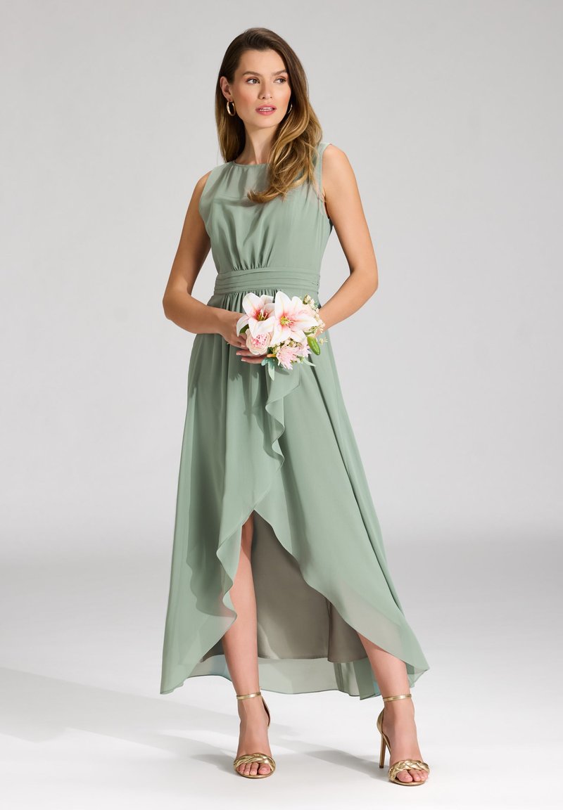 Abito lungo e fluente in chiffon verde chiaro con corsetto aderente, orlo arricciato e design asimmetrico. Indossato con tacchi dorati a strappi e tenendo dei fiori.