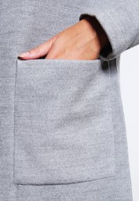 Cappotto di lana grigio con tasca quadrata, caratterizzato da una texture liscia e una mano visibile che si infila nella tasca.