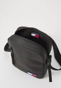 Black crossbody bag made of smooth material, featuring a zippered compartment and a fabric logo patch with red, white, and blue accents.