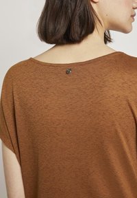 Hnědý top s krátkým rukávem a texturovaným povrchem, který má široký výstřih a dekorativní knoflík na zadní straně. Měkká látka s volným střihem.