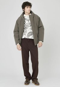Veste rembourrée olive avec capuche au-dessus d'un pull blanc à motif paisley noir, assortie à un pantalon marron et des chaussures marron à lacets.