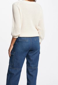 Cremefarbener Strickpullover mit Dreiviertelärmeln, kombiniert mit dunkelblauen Cargo-Jeans mit seitlichen Taschen und detaillierter Naht.