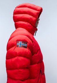 Chaqueta acolchada roja con una gran capucha, que presenta un parche de la bandera noruega en la manga. Textura suave y cierre de cremallera en la parte delantera.