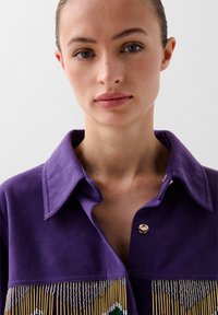 Camicia in suede viola con colletto, caratterizzata da dettagli di frangia in perline dorate metallizzate con accenti verdi e bianchi. Chiusura con bottoni al collo.