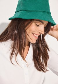 Groene corduroy emmertje met een brede rand. Soepele textuur en eenvoudig ontwerp. Model draagt een witte blouse eronder.