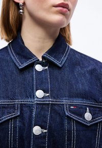 Gros plan d'une personne portant une veste en jean bleu foncé avec des coutures blanches, des boutons métalliques et un petit écusson logo rouge, blanc et bleu.
