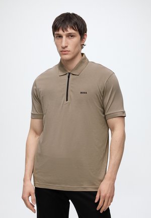 Jeune homme portant un polo beige à manches courtes avec une fermeture éclair à l'avant et le logo "BOSS" sur le côté gauche de la poitrine, debout devant un fond blanc uni.
